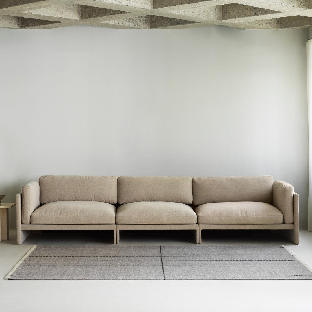 Beige modern sofa with plush cushions, set against a soft grey wall and a textured grey rug. Minimalist design.