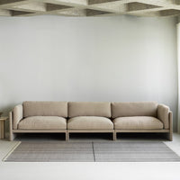 Beige modern sofa with plush cushions, set against a soft grey wall and a textured grey rug. Minimalist design.