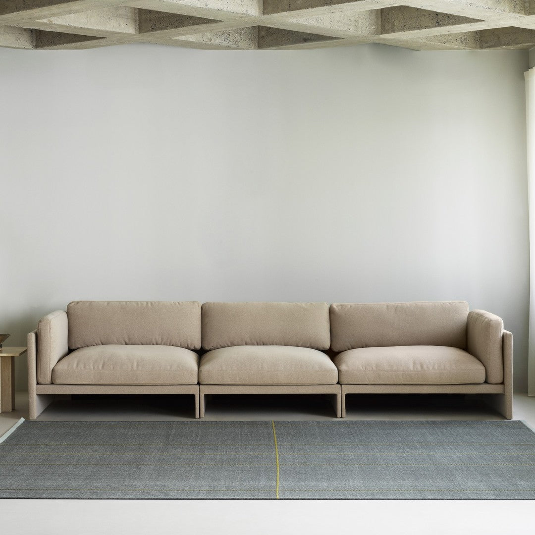 Beige modern sofa on a gray rug with subtle yellow lines against a minimalist backdrop. 