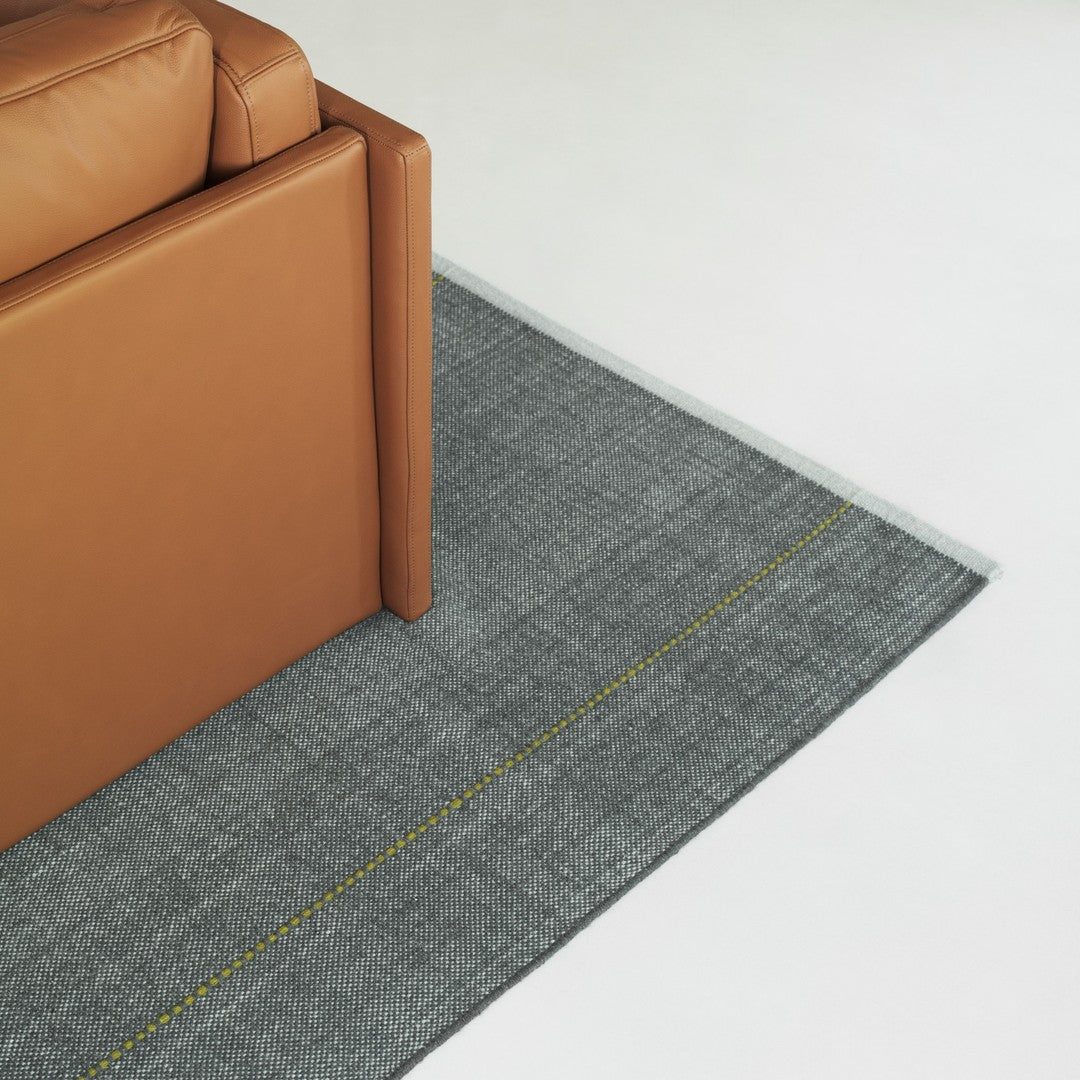 Gray rug featuring a textured pattern and a bright yellow line, complemented by a tan leather sofa edge.