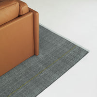 Gray rug featuring a textured pattern and a bright yellow line, complemented by a tan leather sofa edge.