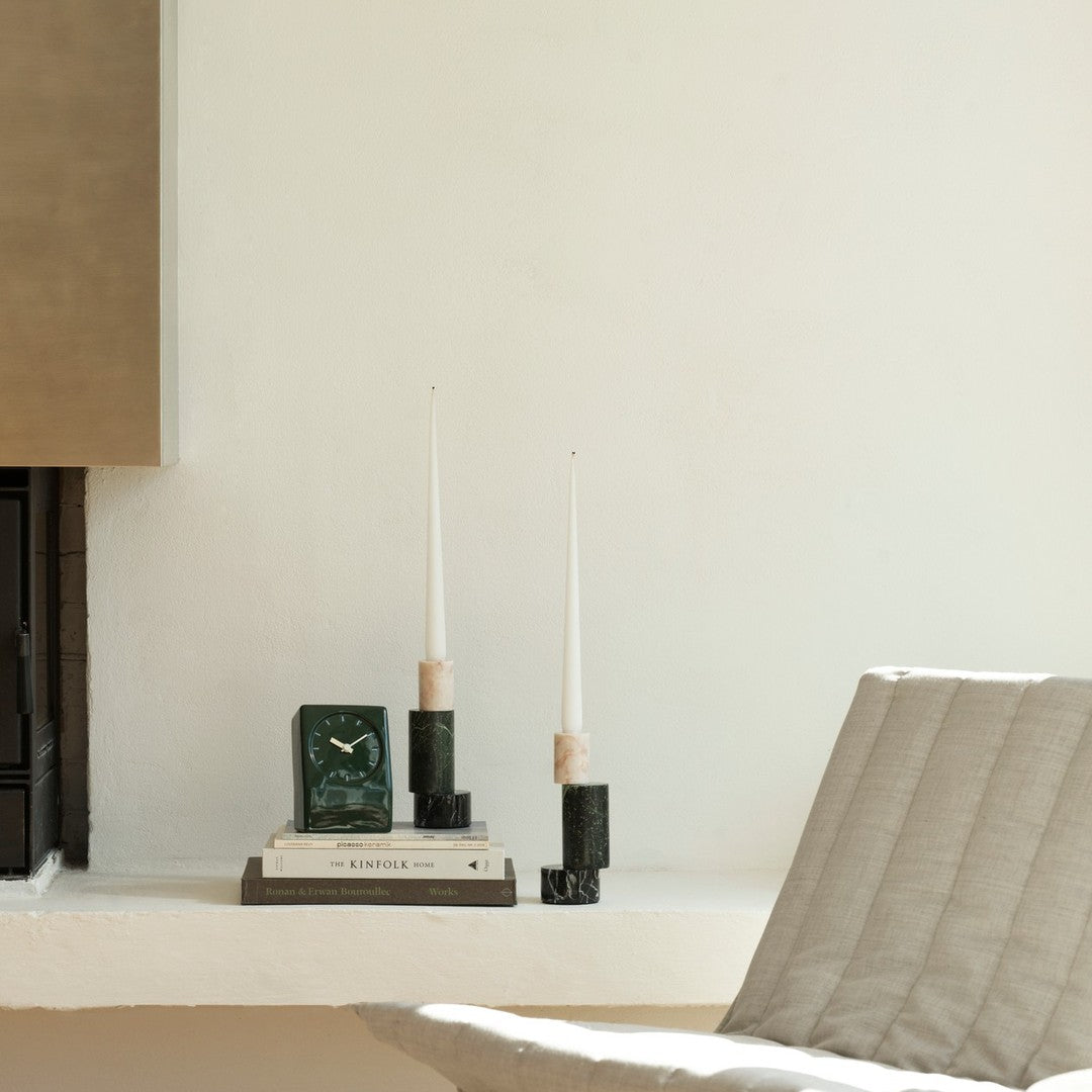 Elegant green and black candle holders with white candles, stacked books, and a textured cream chair. Minimalist design.