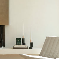 Elegant green and black candle holders with white candles, stacked books, and a textured cream chair. Minimalist design.
