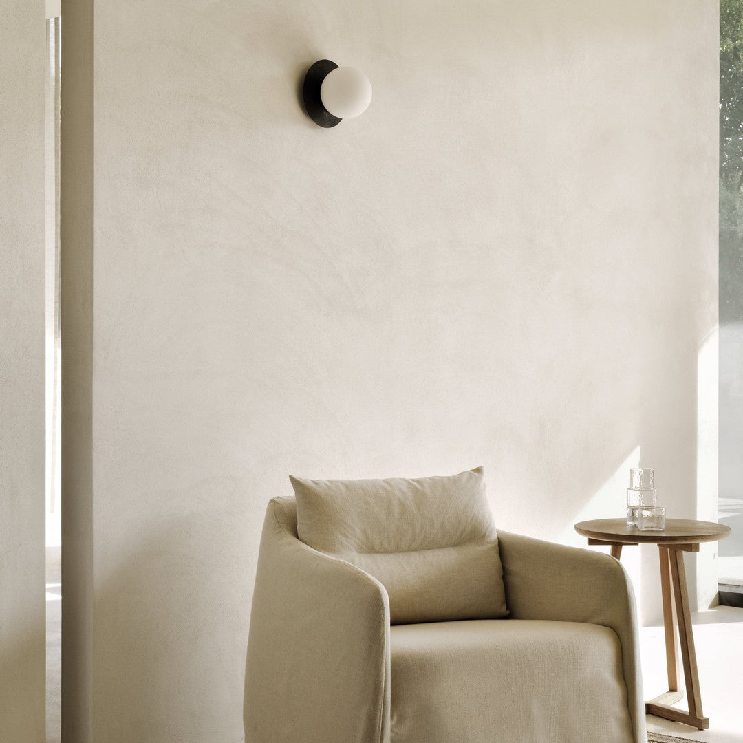 Beige fabric armchair with a smooth texture against a soft, neutral-toned wall, complemented by a black and white lamp.