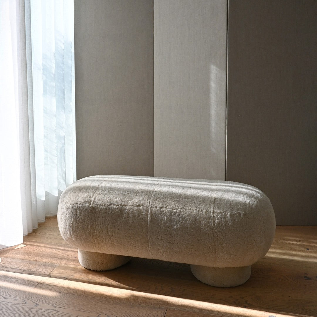 Beige, textured upholstery on a smooth, oval-shaped bench against neutral backdrop with wooden floor accents.