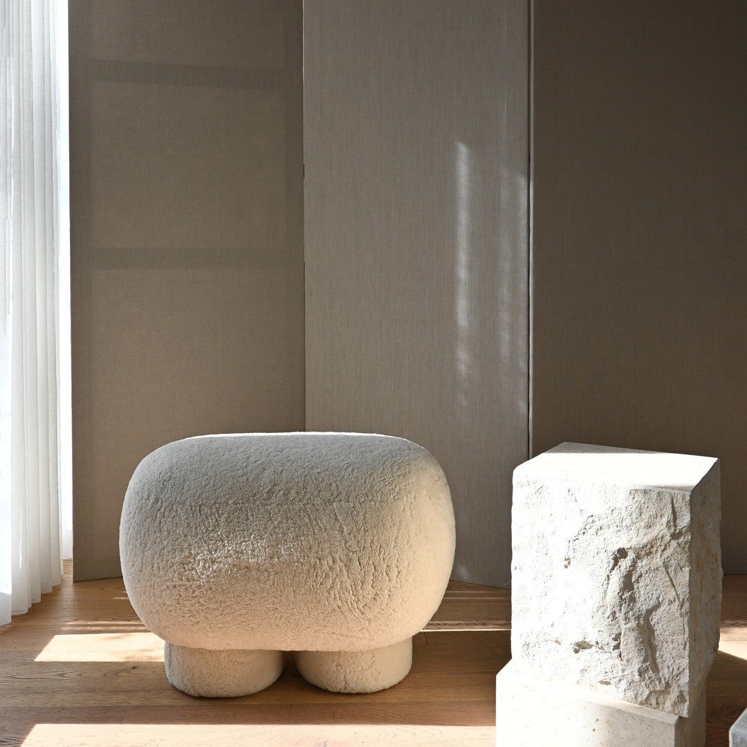 Textured beige ottoman with soft, rounded edges on a light wooden floor, complemented by a smooth white stone pedestal.