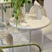 "Round white table with subtle ribbed pattern, paired with cream chairs and green accents, showcasing modern minimalist design."