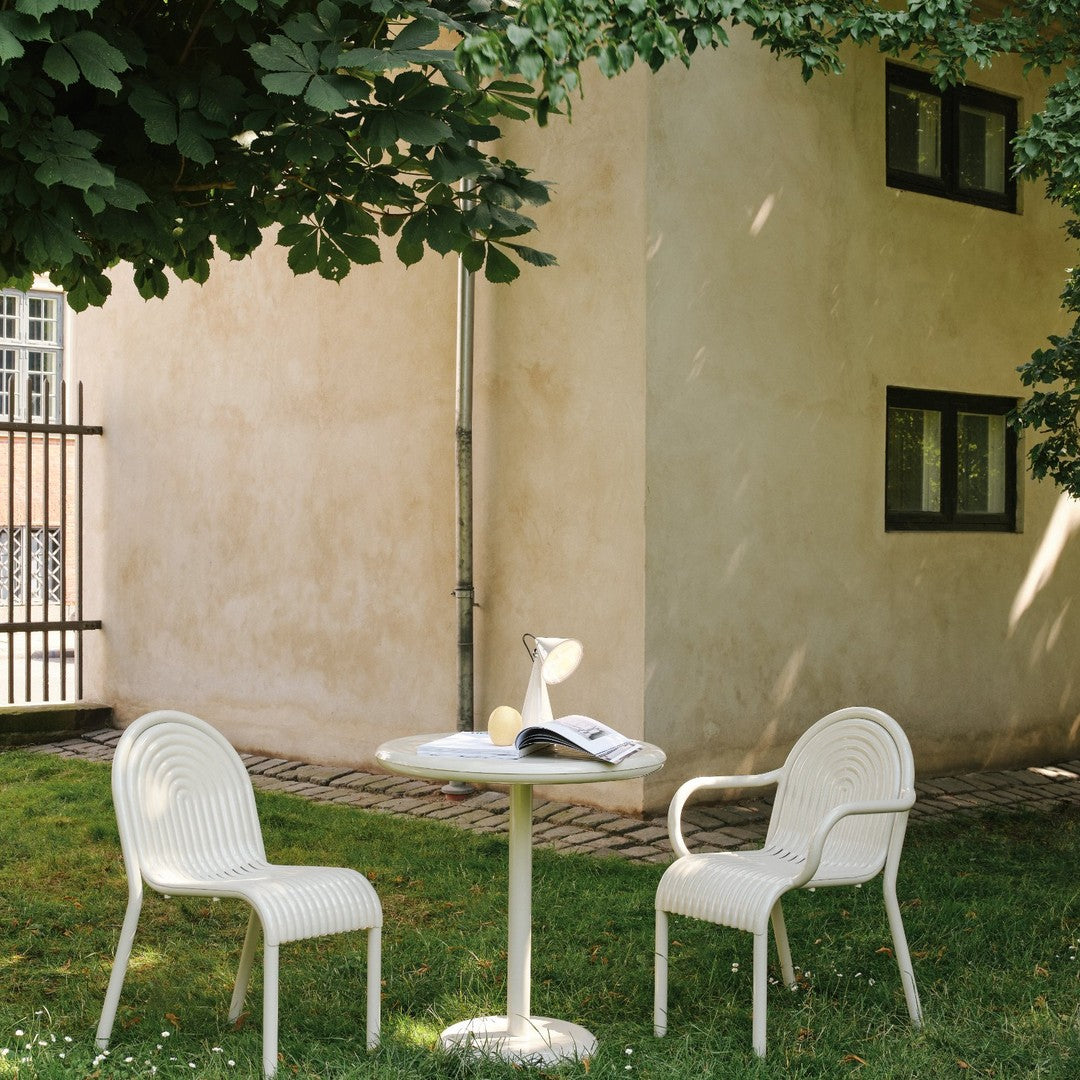 White outdoor table and chairs with subtle ribbed patterns, set against a muted backdrop of beige and green foliage.