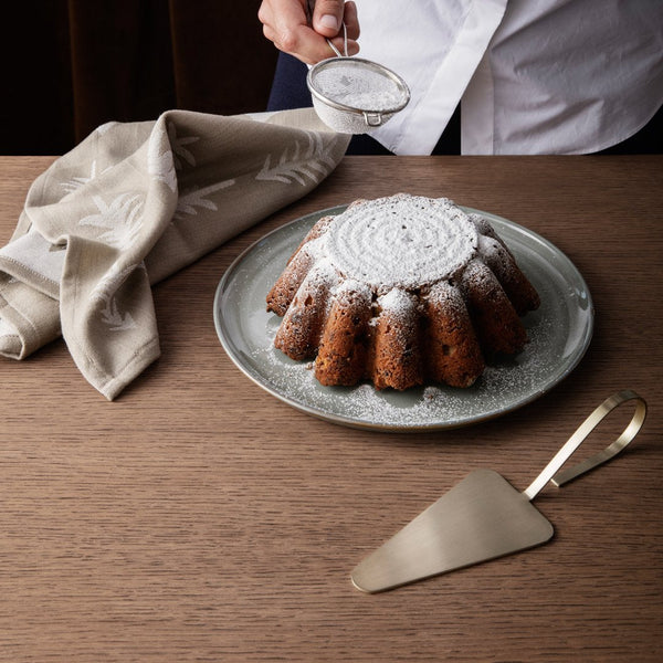 Brown bundt cake dusted with powdered sugar on a gray plate, alongside a beige patterned napkin and gold server.
