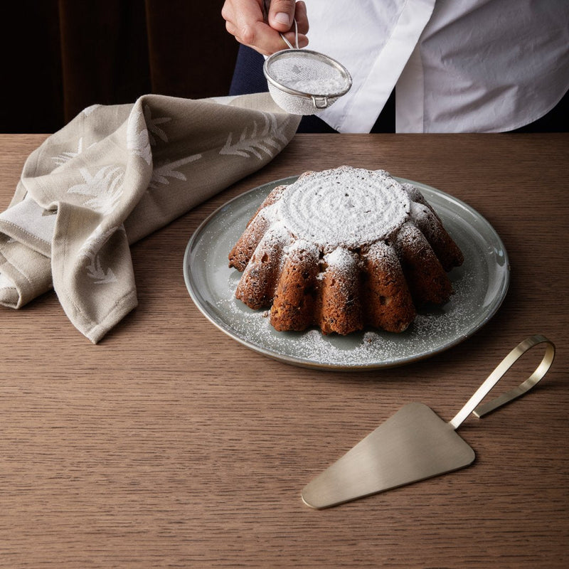 Brown bundt cake dusted with powdered sugar on a gray plate, alongside a beige patterned napkin and gold server.