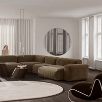 "Curved brown velvet sofa in a minimalistic room with gray walls and a round mirror reflecting light."
