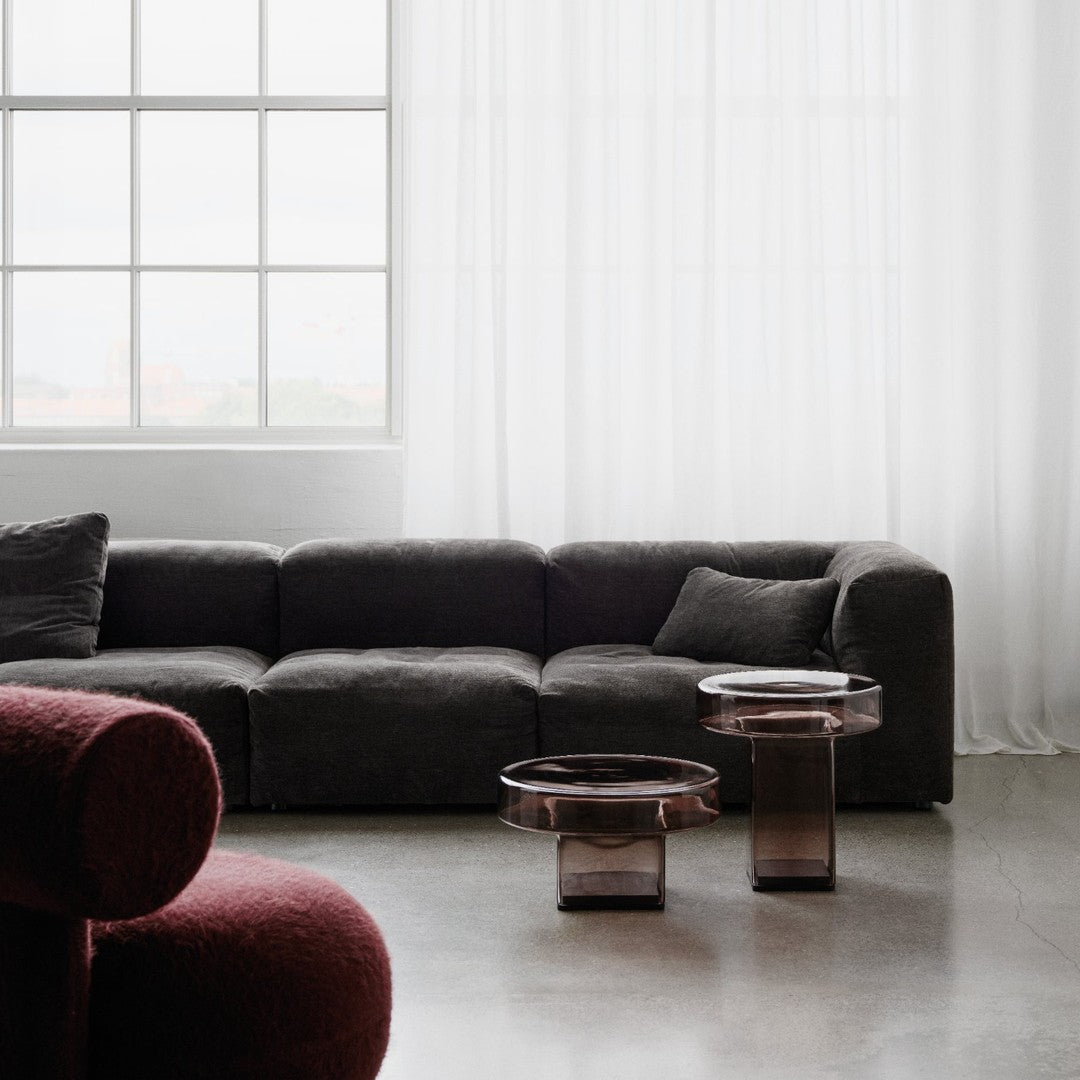 Modern living room with a dark gray sofa and burgundy accent chair, featuring clear and smoky glass tables.