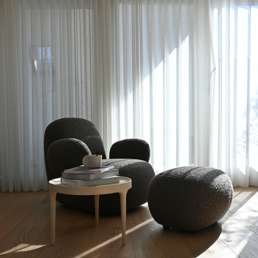 Cozy gray textured chair and ottoman set with light wooden table and white curtains against a sunlit backdrop.