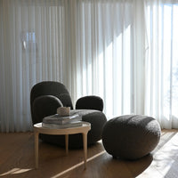 Cozy gray textured chair and ottoman set with light wooden table and white curtains against a sunlit backdrop.