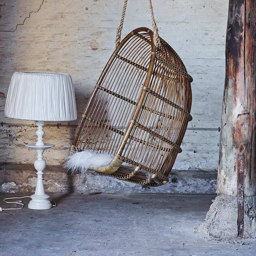 "Woven rattan hanging chair with rustic brown tones, complemented by a white fringed lampshade in an industrial setting."