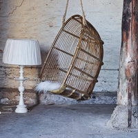 "Woven rattan hanging chair with rustic brown tones, complemented by a white fringed lampshade in an industrial setting."
