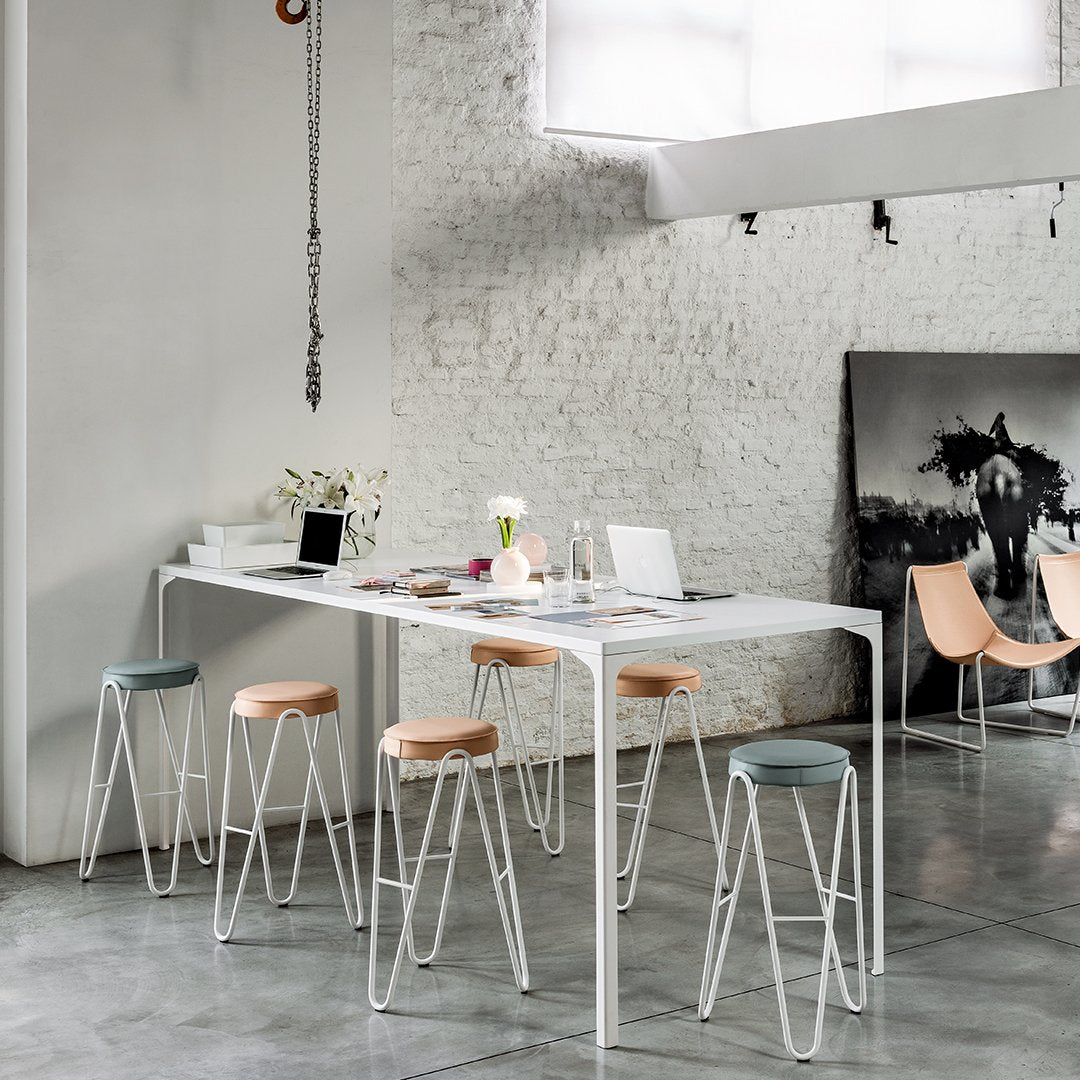 Modern dining room featuring a white table, light wood and pastel stools, with a textured gray floor and brick wall.