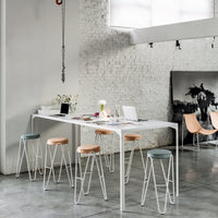 Modern dining room featuring a white table, light wood and pastel stools, with a textured gray floor and brick wall.
