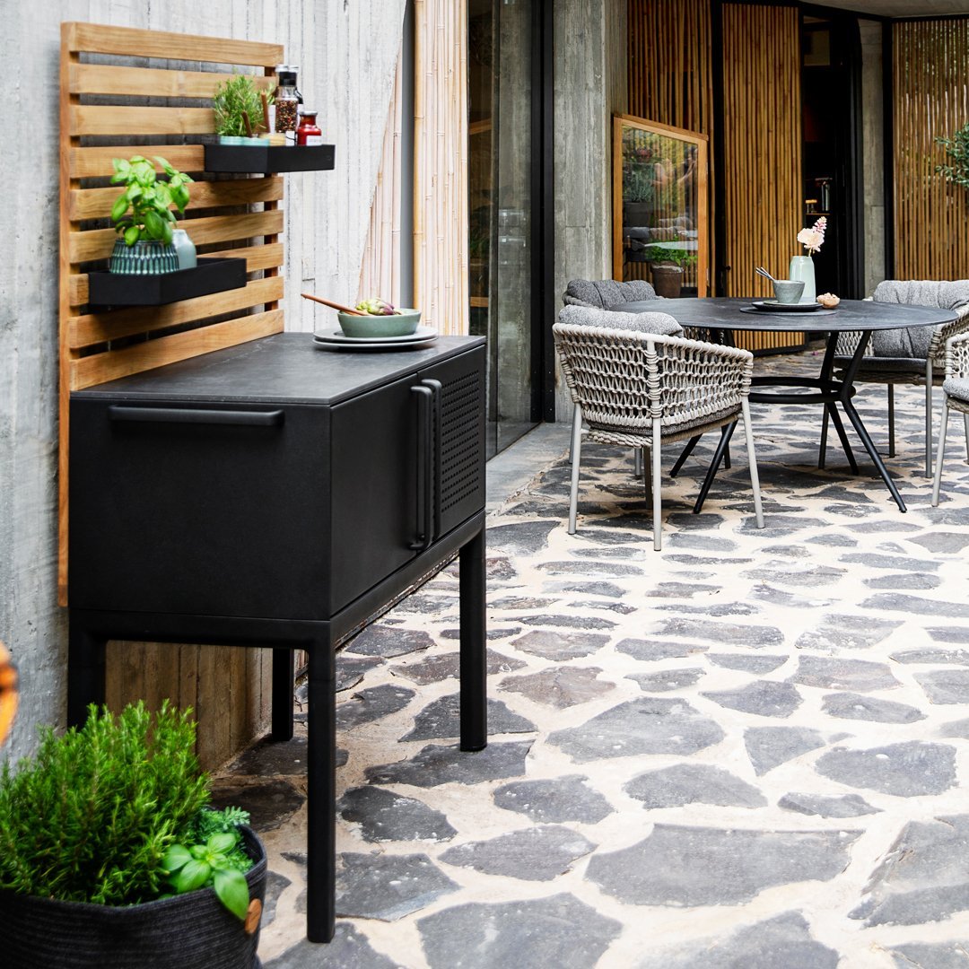 "Black outdoor cabinet on stone-patterned patio, accented by green plants and wooden decor."
