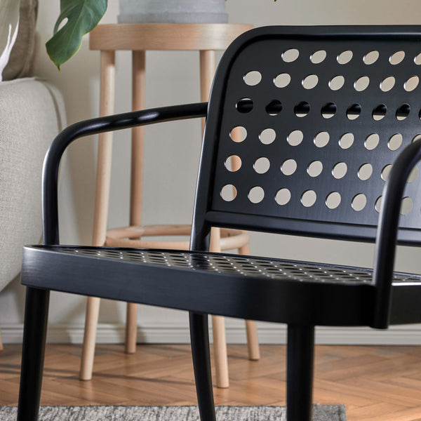 Black metal chair with perforated pattern and round cutouts, set against light wooden accents and a neutral-toned backdrop.