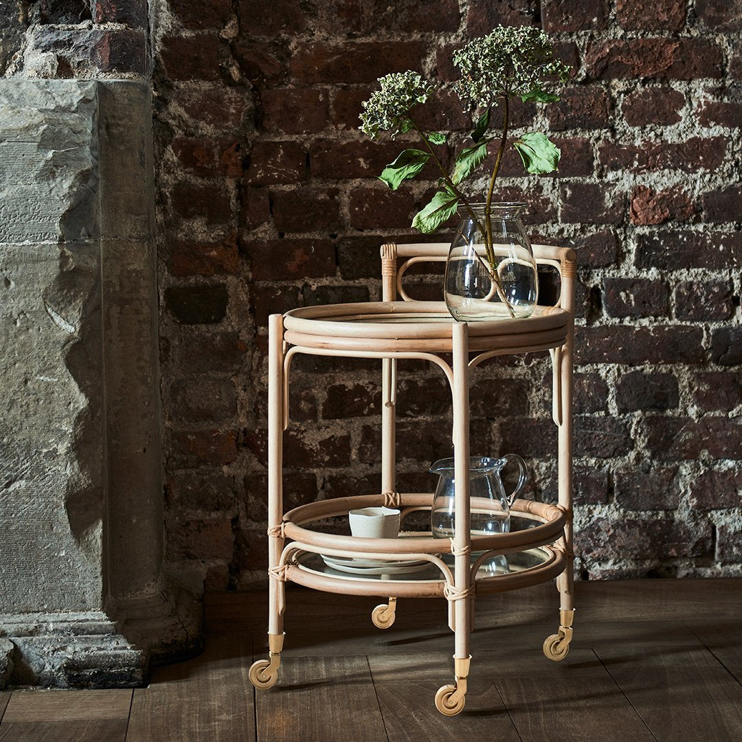 Natural rattan end table with a light wood finish, featuring circular shelves and elegant curved details against a dark brick wall.