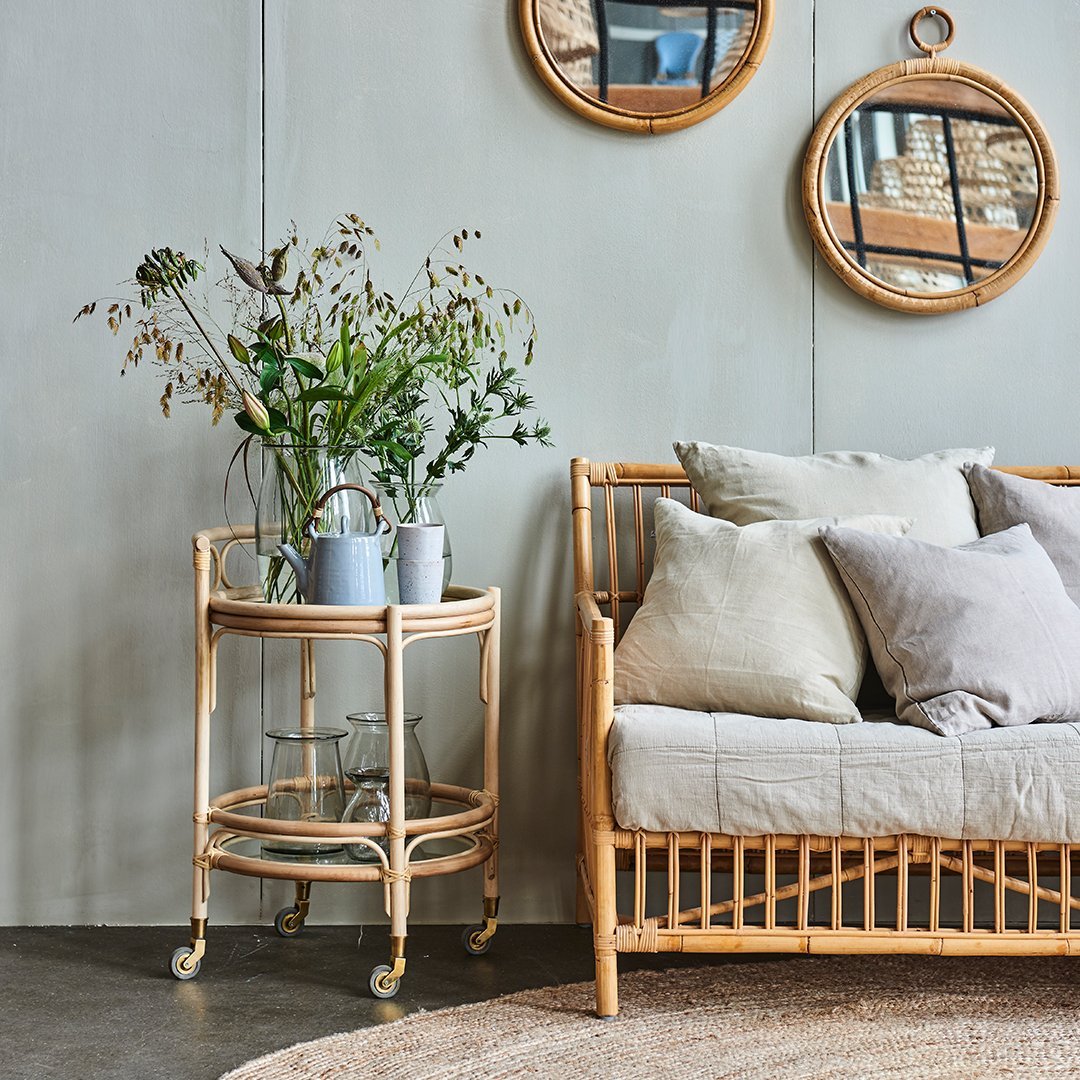 Natural rattan serving cart with a light wood finish, displayed alongside neutral-toned pillows and green floral arrangements.