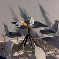 Black square table with modern design, paired with gray chairs on a wooden deck, accented by orange fruit and a silver pitcher.