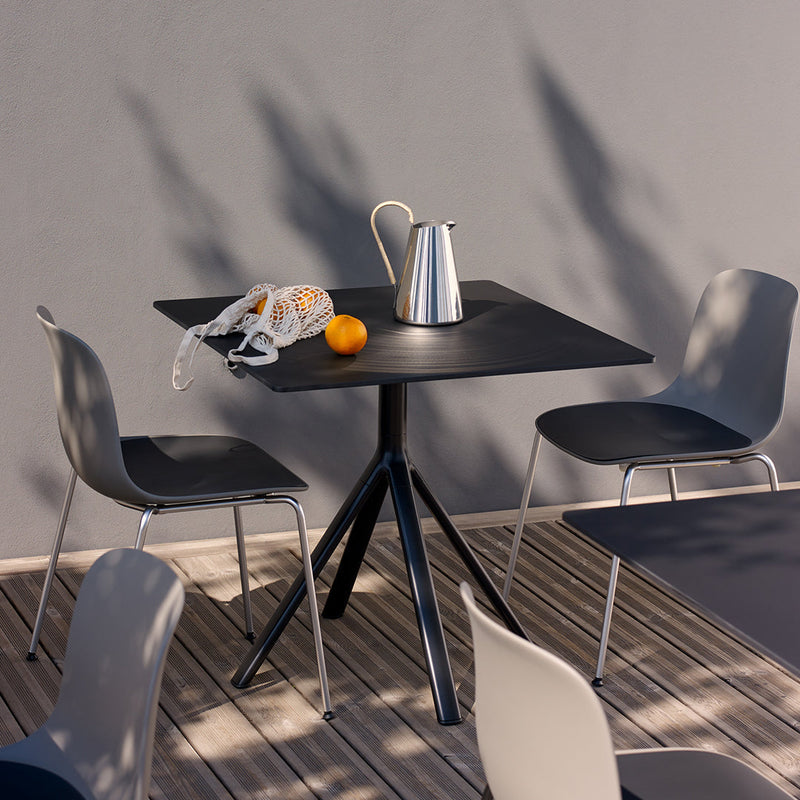 Black square table with modern design, paired with gray chairs on a wooden deck, accented by orange fruit and a silver pitcher.