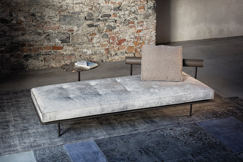Gray velvet chaise lounge with tufted design and a beige cushion, set against a textured stone wall.