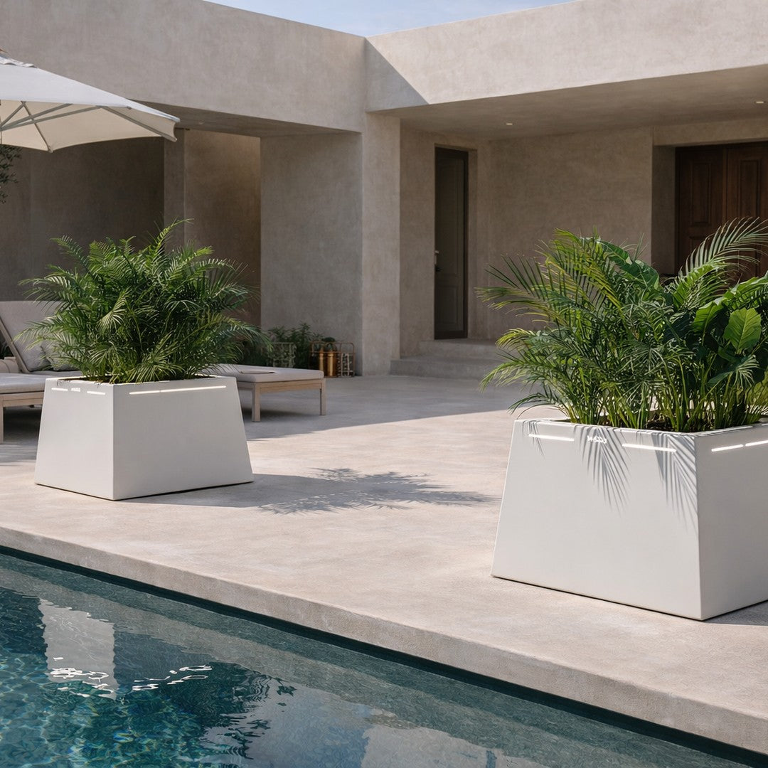 Modern white geometric planters with lush green plants, set against a neutral patio and turquoise pool backdrop.