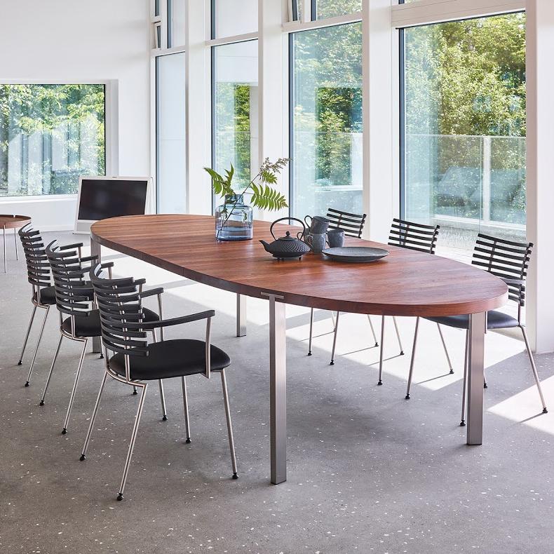 Modern wooden table with a rich brown finish, surrounded by black striped chairs, set in a bright, airy space.