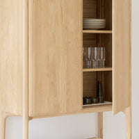 Natural oak cabinet with smooth, light wood grain and minimalist design; open shelves display glassware and plates.
