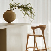Neutral tones featuring a textured beige vase, light wood chair, and soft white curtains with green foliage accents.