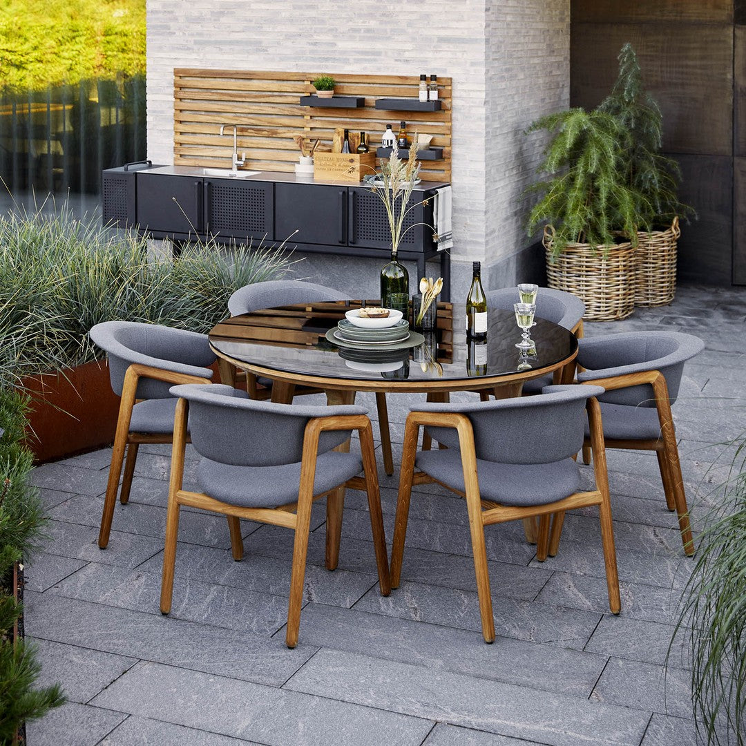 "Round outdoor dining table with a wooden top, gray cushions, and natural wood accents, surrounded by lush greenery."