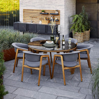 "Round outdoor dining table with a wooden top, gray cushions, and natural wood accents, surrounded by lush greenery."