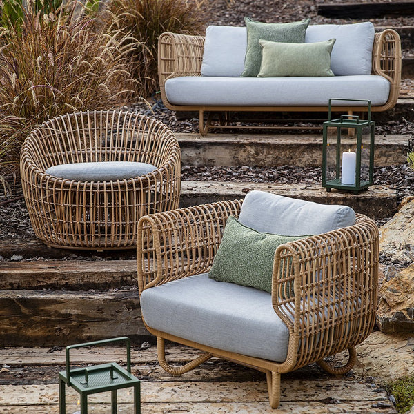 Natural rattan furniture set featuring grey cushions and a green accent pillow, set against a rustic outdoor backdrop.