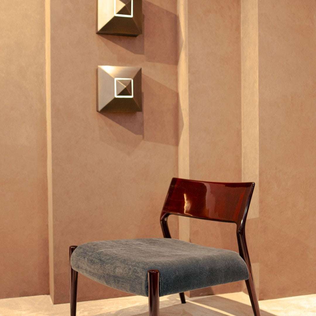 Stylish chair with a dark grey velvet cushion and glossy wooden legs against a warm beige wall, featuring geometric decor.