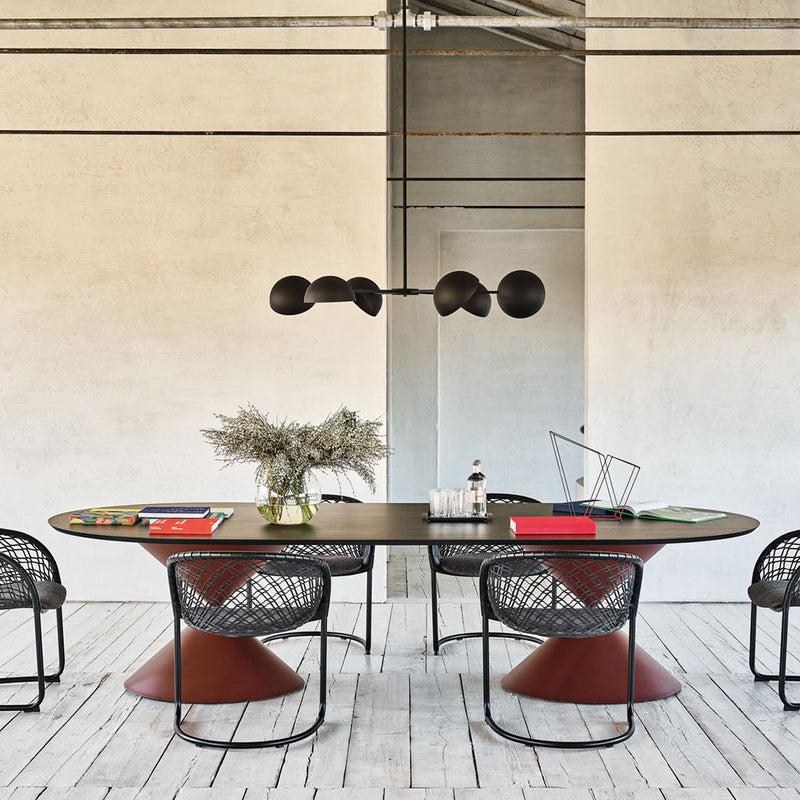 Stylish oval dining table with a sleek black surface and contrasting red cone base, surrounded by modern black mesh chairs.