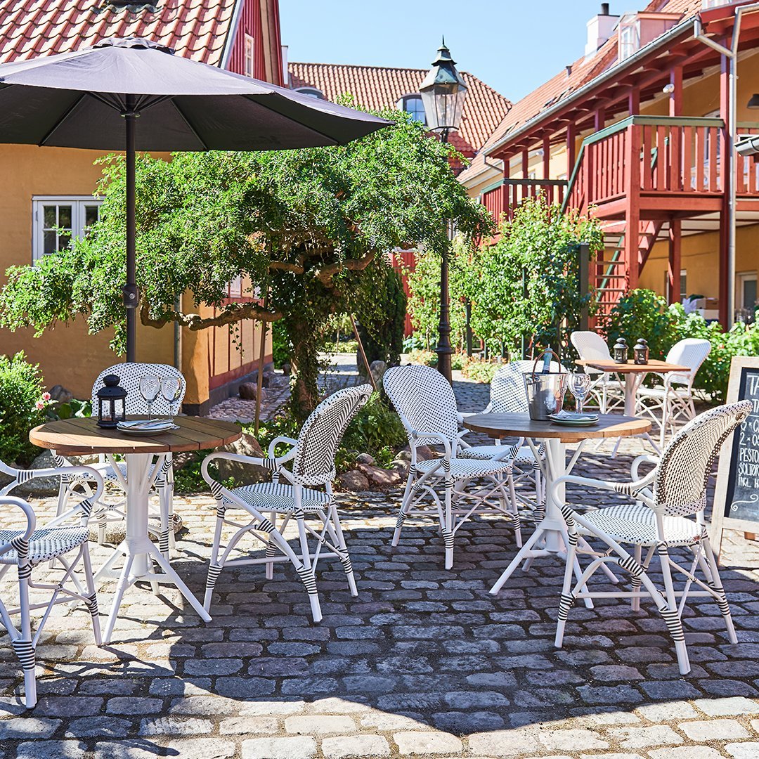 White wicker chairs and round tables set against vibrant red and yellow buildings, with green foliage and a gray cobblestone patio.