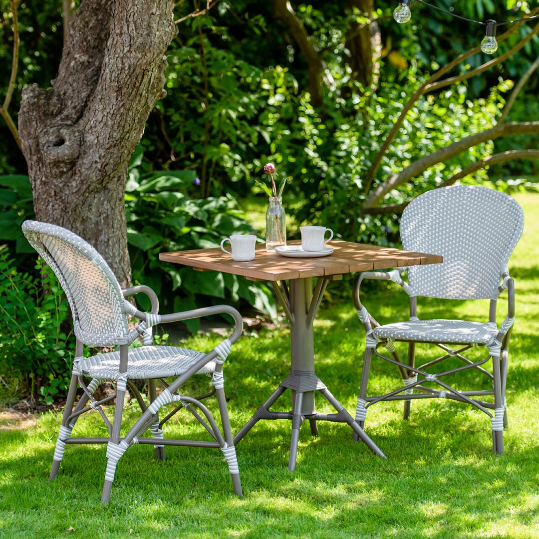 Gray wicker chairs with textured patterns, natural wood table, and white mugs in a lush green garden setting.