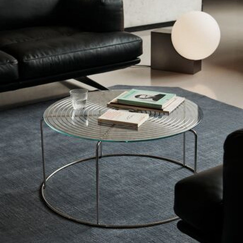 Round glass coffee table with a textured surface, silver metal frame, and placed on a dark blue rug. Modern living room decor.