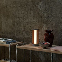 Modern lamp with vertical orange stripes, paired with glossy burgundy vase and ceramic bowl on log shelf.