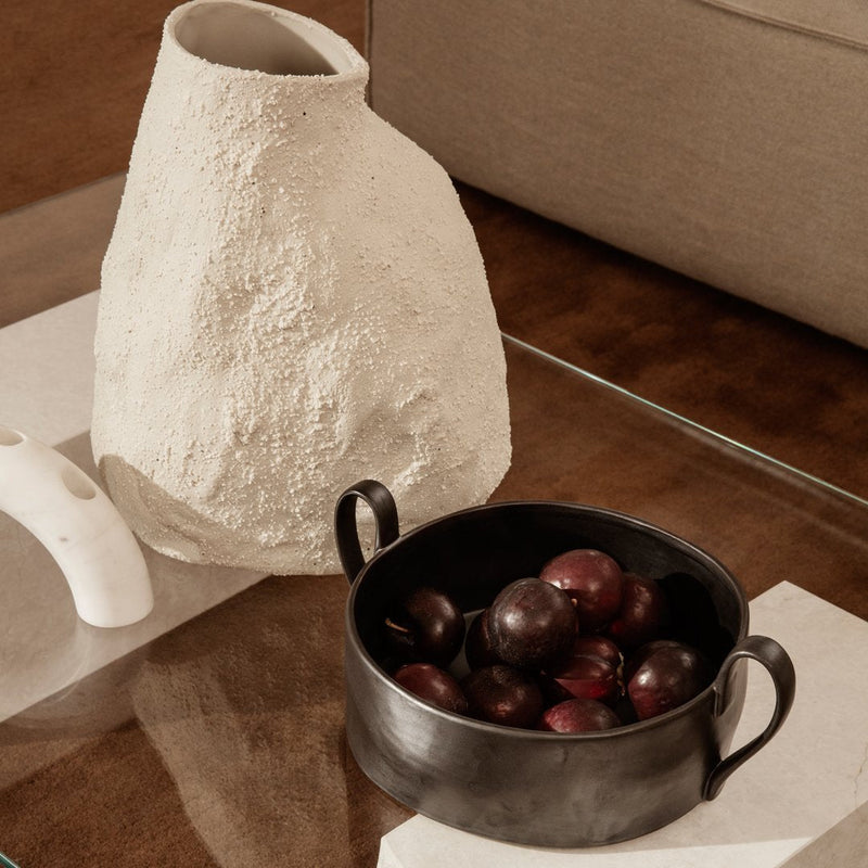 Textured white vase beside a black bowl filled with deep purple fruits, situated on a clear glass table.