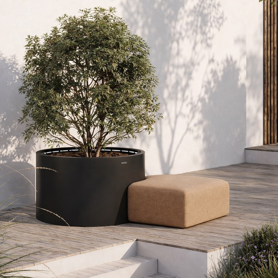 Black round planter with a lush green tree and a tan rectangular stool set against light wooden decking.