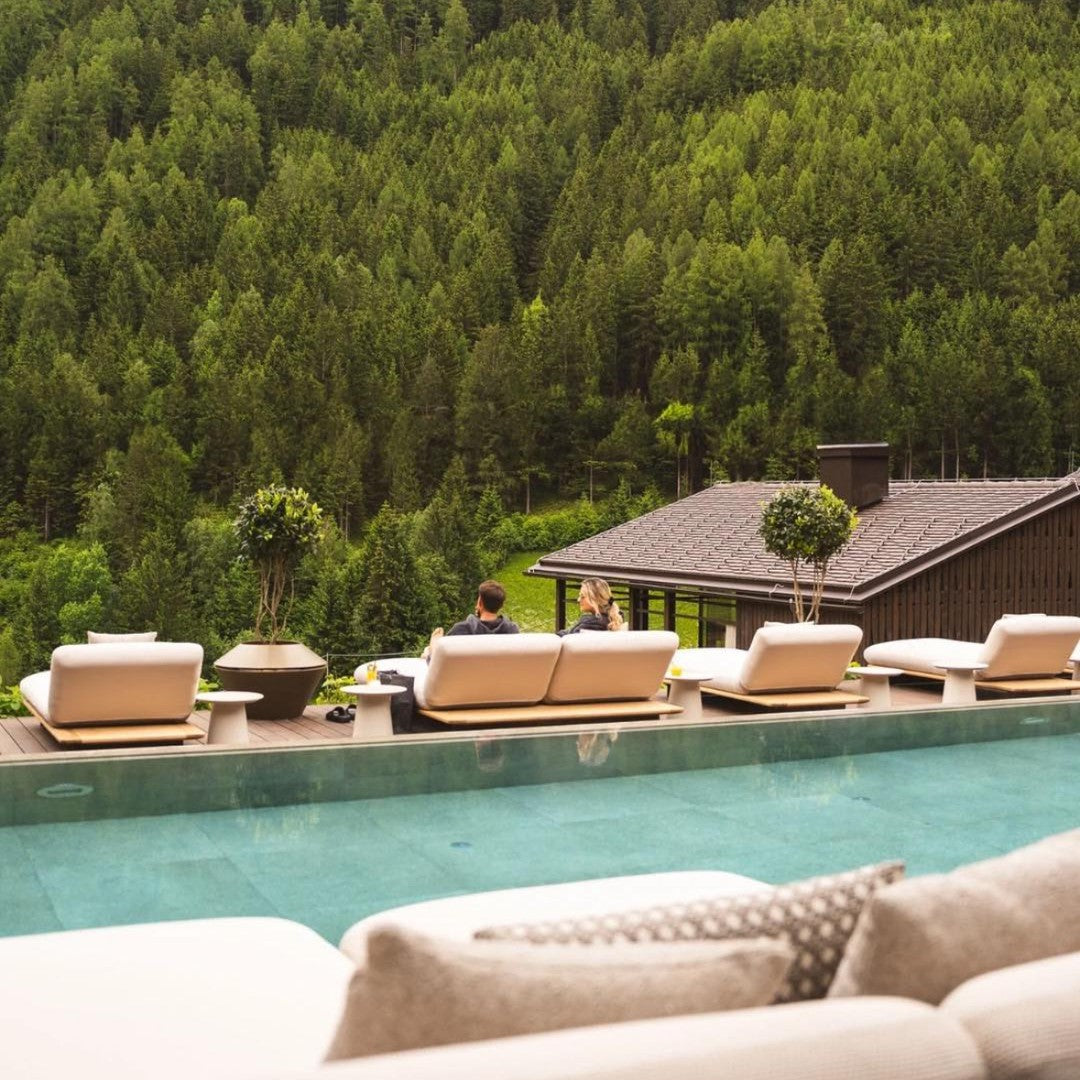 Neutral beige outdoor furniture with green shrub accents, set against a lush forest backdrop and tranquil blue water.