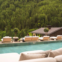 Neutral beige outdoor furniture with green shrub accents, set against a lush forest backdrop and tranquil blue water.