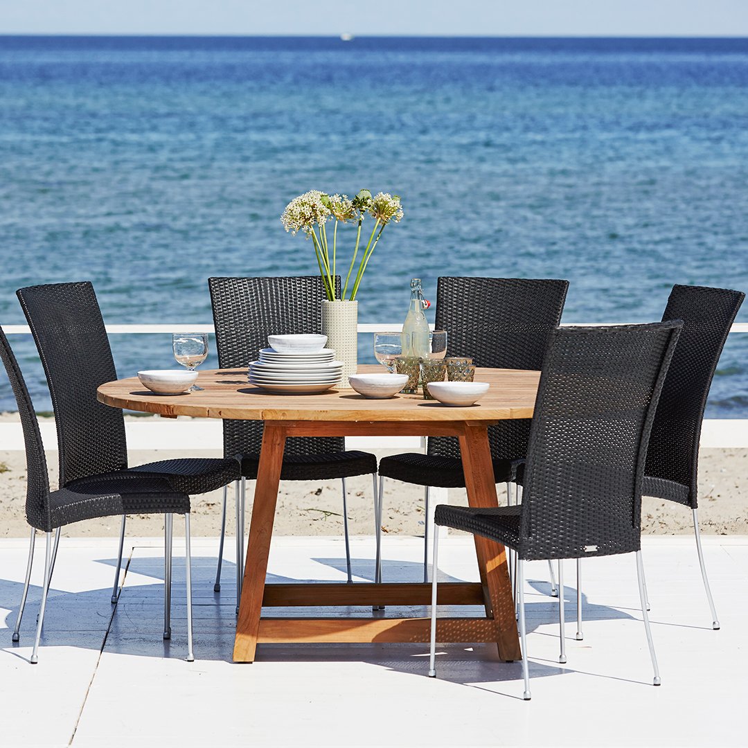 Round wooden patio dining table with six black wicker chairs against a serene blue ocean backdrop.