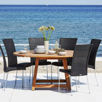 Round wooden patio dining table with six black wicker chairs against a serene blue ocean backdrop.