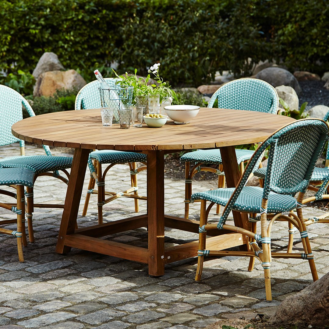 Round wooden dining table with teal wicker chairs, set on a textured stone patio with greenery in the background.