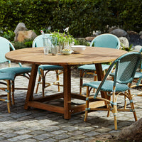 Round wooden dining table with teal wicker chairs, set on a textured stone patio with greenery in the background.